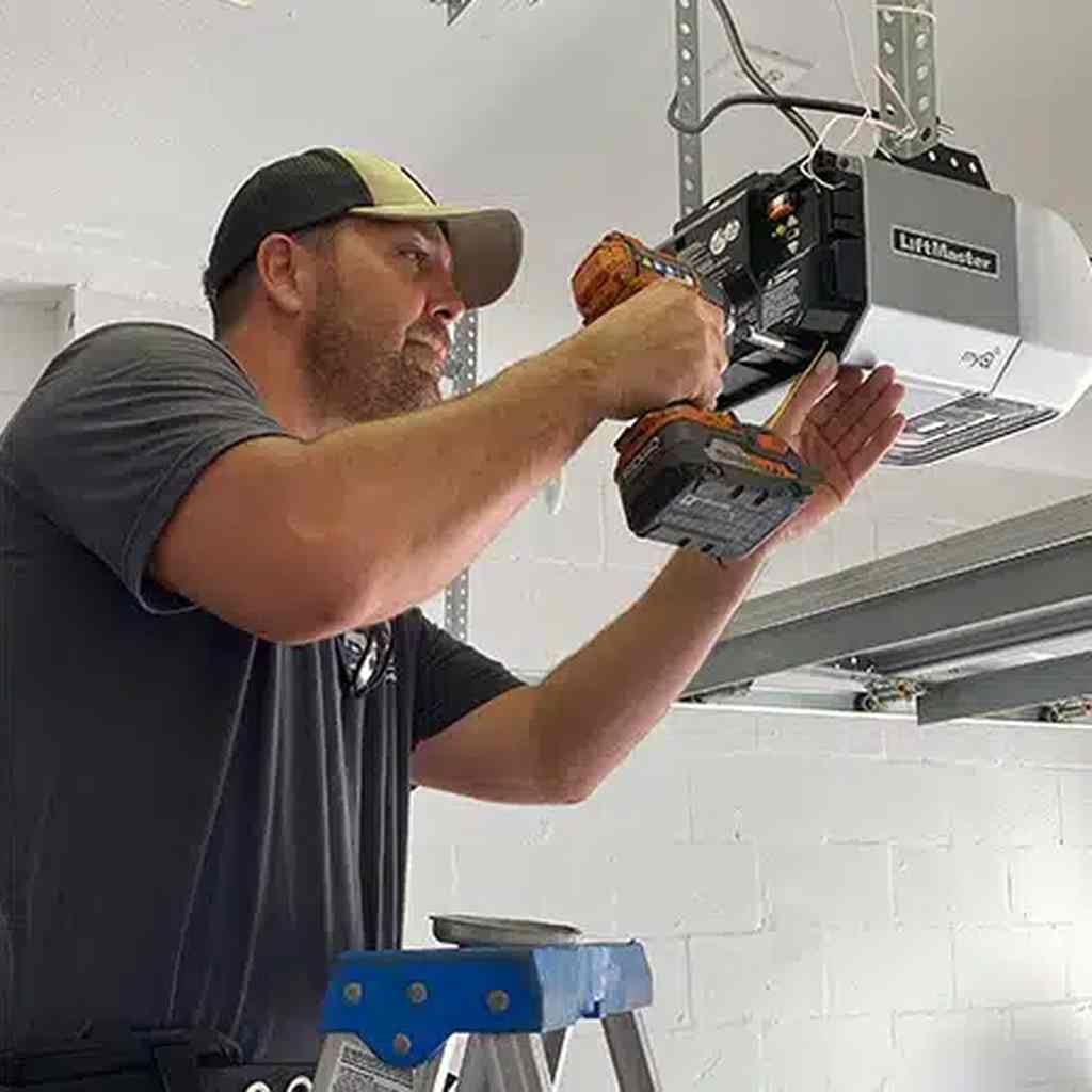 Bearded technician in a dark gray t-shirt and two-tone cap using a cordless drill to install or service a LiftMaster garage door opener while standing on a blue step ladder.